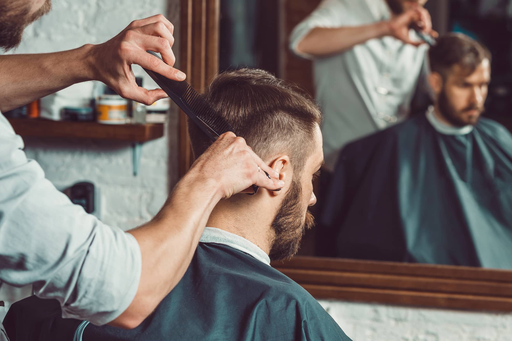 As mãos do jovem barbeiro fazendo corte de cabelo de homem barbudo atraente na barbearia